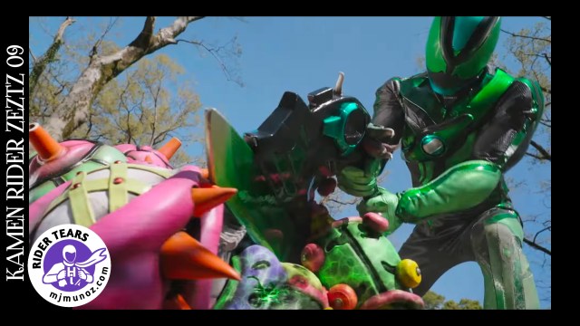 Kamen Rider Zeztz in his green-and-black axe form swings his massive weapon down at a grotesque, vegetable-like monster covered in colorful spikes. The scene is full of vibrant motion and tension, framed by bright daylight and the bare limbs of tall trees.