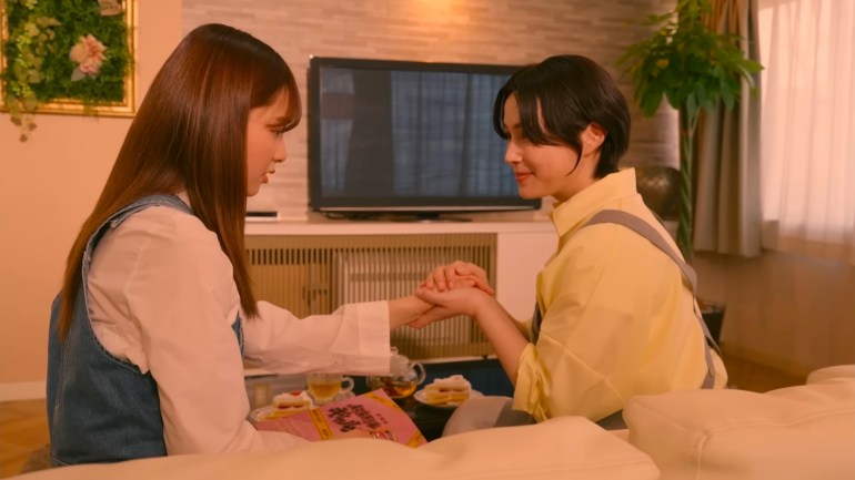 The Lady and Nem sit facing each other on a couch, holding hands in a warmly lit living room.