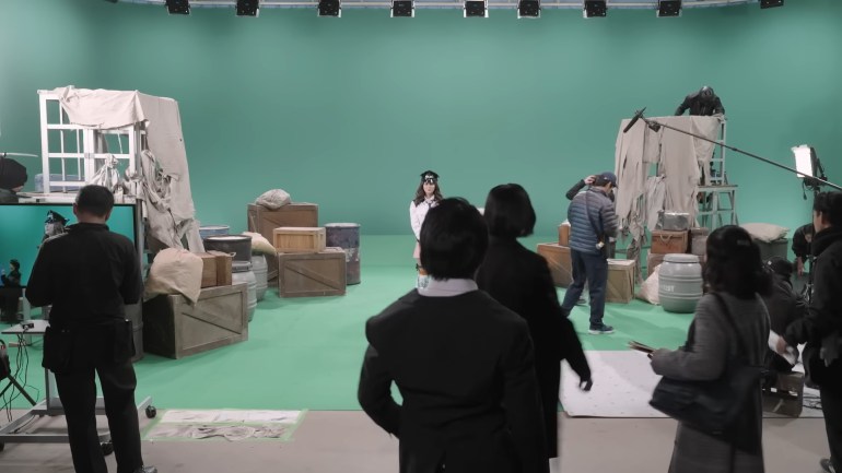 A large green screen studio with prop crates and barrels, crew members, lighting equipment, and a monster performer standing on an elevated platform while a scene is prepared.