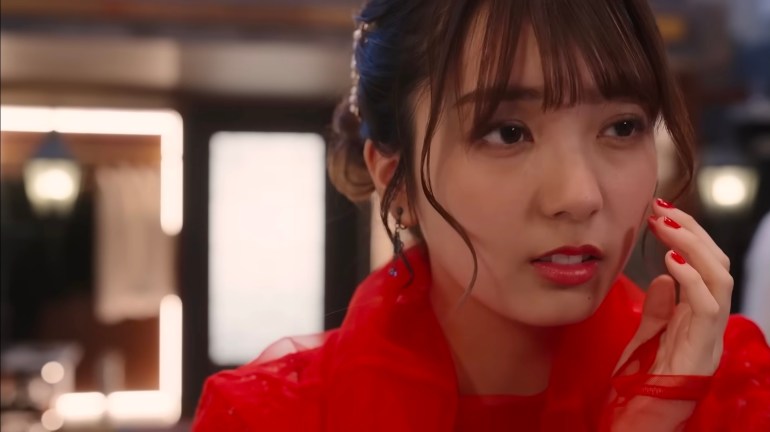 Nem, wearing a bright red outfit, touches her face while looking off to the side.