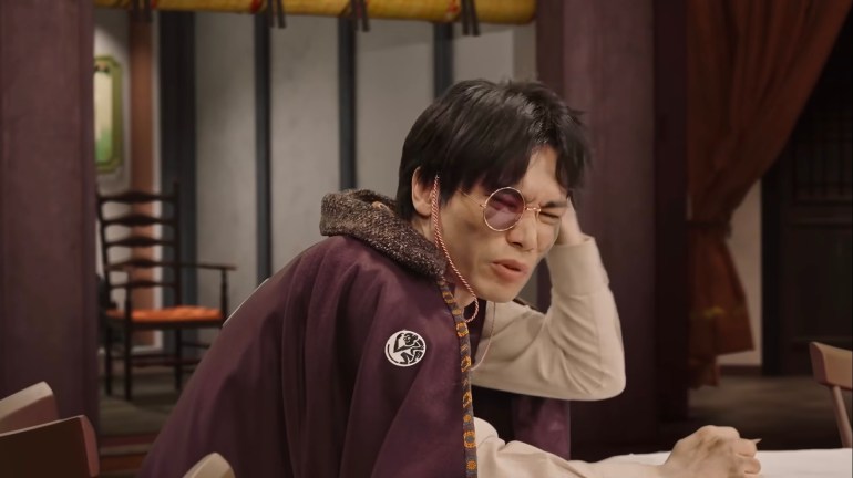 Ende sits at a table wearing a monocle and holding his head with one hand while looking frustrated and confused.