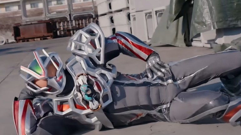 Kamen Rider My-Th reclines on the ground in silver, gray, and red armor, holding one bent leg while extending the other.