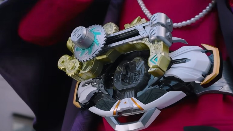 Close-up of a silver and gold mechanical device featuring exposed gears, a sliding mechanism, turquoise markings, and white armor-like panels against a red background.