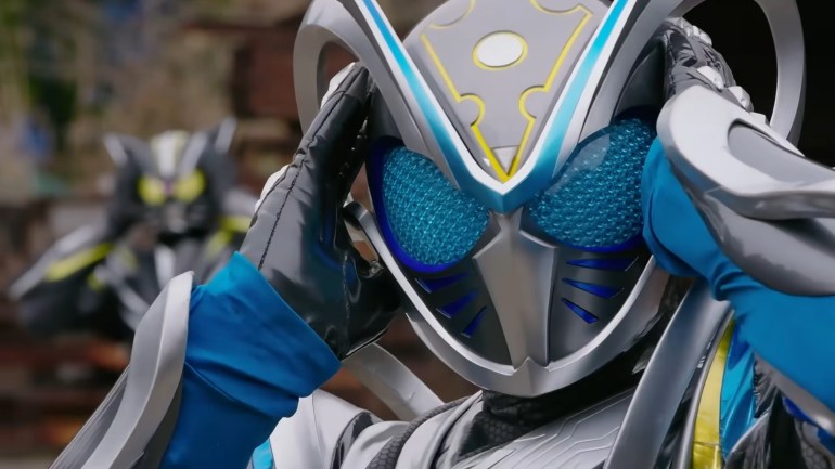 Kamen Rider MY-TH holds both hands against the large ears on his helmet while Kamen Rider Rid stands blurred in the background.
