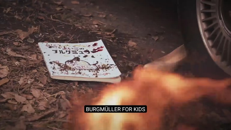 A blood-stained Japanese piano book titled Burgmüller for Kids lies on the ground beside a car wheel and burning wreckage.