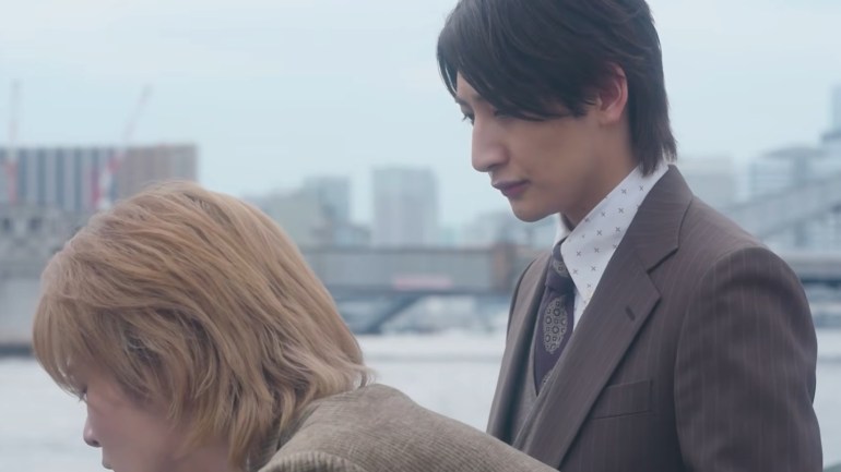 Asao, with brown hair in a suit, stands beside Tigre, who has blond hair, outdoors against a pale city background.
