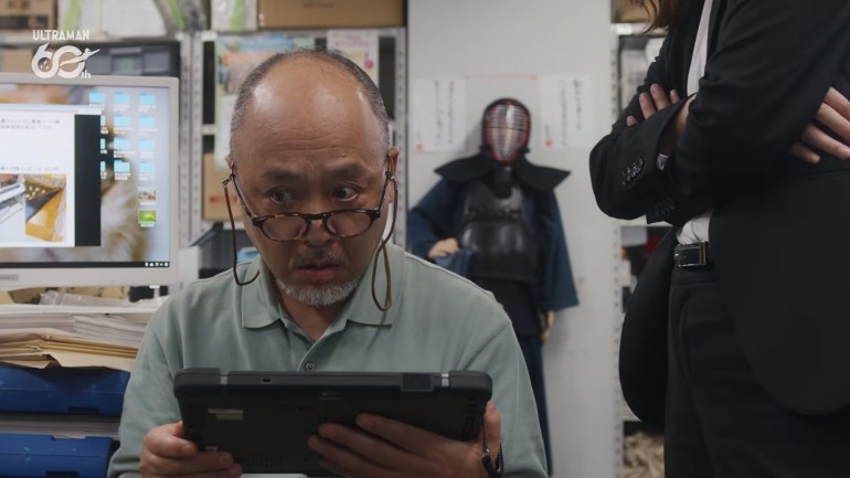 Professor Minobe, wearing glasses and a light green polo shirt, looks nervously to his right while holding a black tablet. A person in a black suit stands beside him with arms crossed.