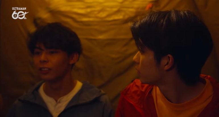 Rintaro, wearing a gray jacket over a white shirt, talks beside Ibuki, who wears an orange jacket and yellow shirt, inside a warmly lit camping tent.