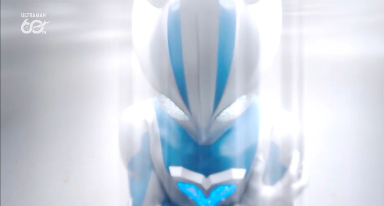 Close-up of Ultraman Teo with his head lowered and both hands raised near his face, surrounded by an intense white glow.