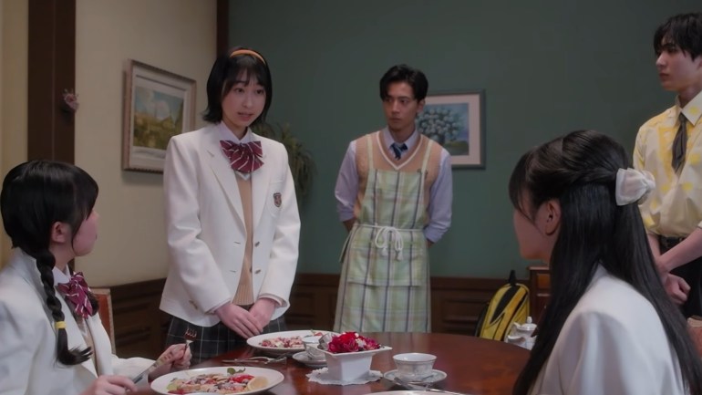 A schoolgirl in a white blazer stands beside a dining table while two other schoolgirls sit nearby. Behind her stands a young man wearing a plaid apron, with another young man in a yellow patterned shirt at the right.