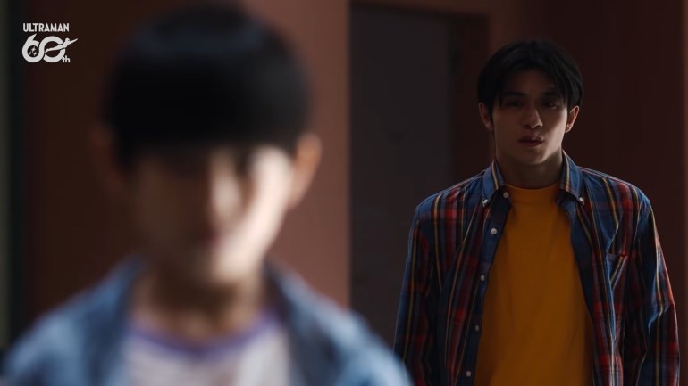 Ibuki, wearing a yellow shirt and a blue-and-red plaid overshirt, faces a young boy who is blurred in the foreground of an indoor scene.