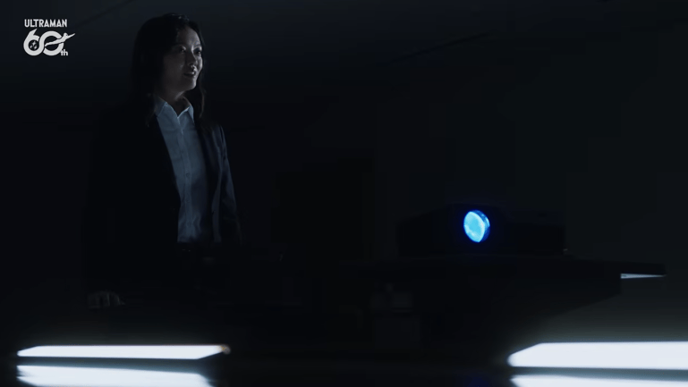 Kyoko Shiga stands in a dark room wearing a black blazer and white shirt, with an incredulous expression on her face. A projector glows blue beside her.